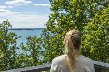 Fototapeta premium Beautiful summer Baltic sea landscape view. Young blond woman looking on sea through green tree tops. 