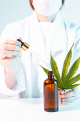 Close up of doctor in mask and robe with a hemp leaf, pipette and mock up bottle of cannabis oil product