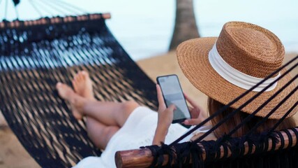 Young woman traveler lying on a hammock and using smartphone at the beach while traveling for summer vacation