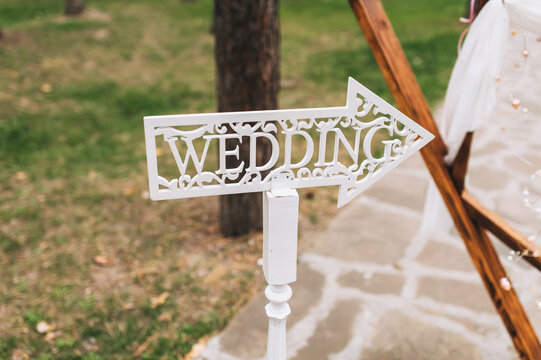 A white plate in the form of an arrow and a pointer with the inscription and letters wedding in English stands in nature at the ceremony. Photography, concept.