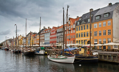 Many boats moored to the dock in the harbor Nyhavn and colorful houses on the embankment against cloudy sky in Copenhagen, Denmark.