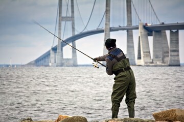 Closeup of a fisherman on the stony coast and Great Belt Fixed link in the background in...