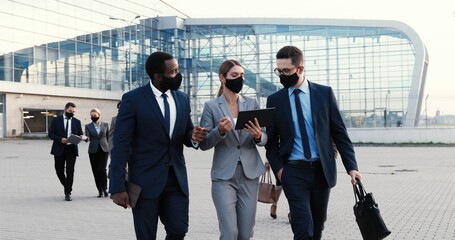 Caucasian businesswoman and two mixed-races businessmen in masks walking outdoor at business center and talking with tablet device in hands. Multi ethnic work partners discussing project and finance.