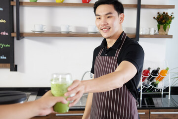 Smiling Asian barista young man serving a plastic glass of iced Matcha green tea and take away to home for customers at counter bar in coffee shop. Start up for bussiness Concept