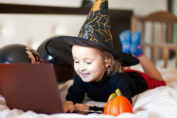 funny child girl in witch costume for Halloween lies on the bed and using a digital tablet laptop notebook. Online call friends or parents