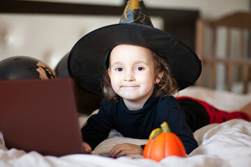 funny child girl in witch costume for Halloween lies on the bed and using a digital tablet laptop...