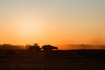 Silhouette of working tractor in autumn field in sunset time, tractor packing hay into rectangular bales, working machine in golden sky background, agriculture concept