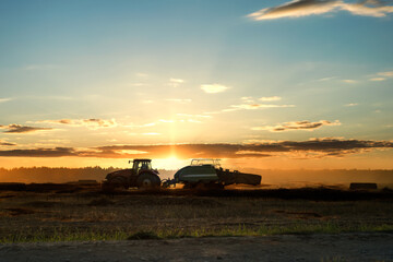 Silhouette of working tractor in autumn field in sunset time, tractor packing hay into rectangular...