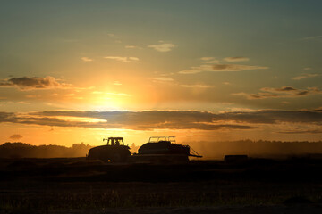 Silhouette of working tractor in autumn field in sunset time, tractor packing hay into rectangular...