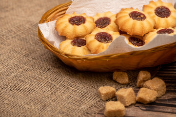 Homemade pastry cookies with jam in a wicker basket and pieces of brown cane sugar on a background of homespun fabric with a rough texture, close-up, selective focus.