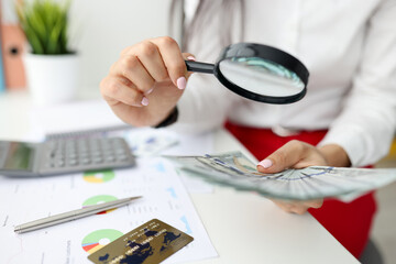 Woman check currency for authenticity. Female hand hold magnifying glass and lot of money. On white table lies gray calculator, metal pen, bank card and document with diagram.