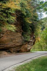 Fall colored trees and shrubs overhang a beautiful stone faced cliff that further overhangs a scenic road through the hill country of Southeast Ohio