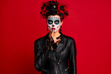 Scary lady calavera. wears artistic make-up for the feast of all the dead. Makes silence gesture keeps finger over lips, wears black leather jacket and lace gloves,dressed as skeleton isolated in red.