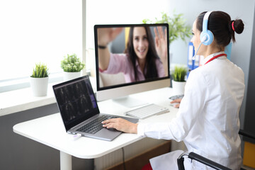 Female doctor in white coat and headphones conduct online consultation. Woman sit at table and work at laptop and computer.