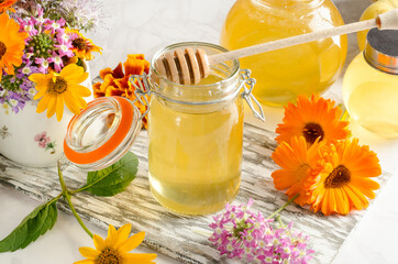 Honey in a glass jar with a honey stick and flowers on a light background