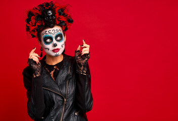 Woman with sugar skull makeup, wreath of flowers on her head and skull and black gloves looks surprised at the free space on the background. isolated on red. concept of Halloween or Calavera Catrina