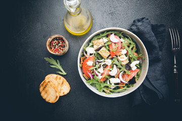 Eggplant salad with tomatoes, arugula and feta cheese in a bowl on black background, top view