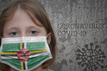 Little girl in medical mask with flag of dominica stands near the old vintage wall with text coronavirus, covid, and virus picture. Stop virus concept