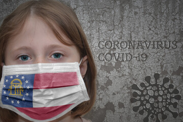Little girl in medical mask with georgia state flag stands near the old vintage wall with text coronavirus, covid, and virus picture. Stop virus concept