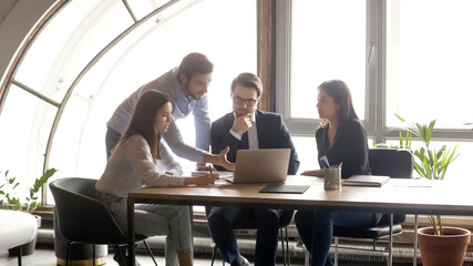Pensive diverse business people teammates employees looking at laptop screen, developing sales growth marketing strategy or analyzing project financial results, brainstorming together in modern office