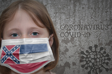 Little girl in medical mask with mississippi state flag stands near the old vintage wall with text coronavirus, covid, and virus picture. Stop virus concept