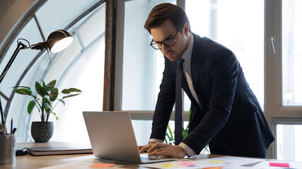 Focused 30s male ceo executive in eyeglasses and formal wear using computer application, preparing...