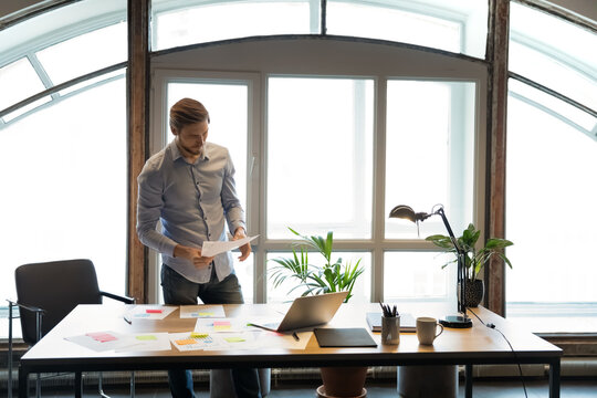 Focused young european manager standing at office table, full of paper document with colorful memo stickers, reading financial or marketing report, analyzing sales statistics brainstorming indoors.