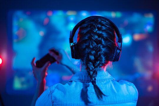 Close-up view from back of gamer girl playing video game at home in front of big screen with joystick and headphone. Colorful neon led lights background. Streamer concept.