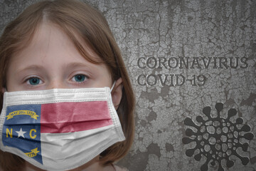 Little girl in medical mask with north carolina state flag stands near the old vintage wall with text coronavirus, covid, and virus picture. Stop virus concept