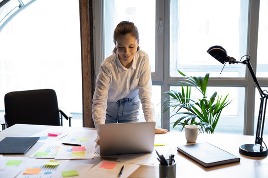 Smiling young european appearance female worker standing at office table, using computer application planning current issues or problems solution, managing tasks with stickers on paper documents.
