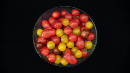
Small cocktail tomatoes, multicolored, round and oval, in a transparent glass container, on black background, close-up	