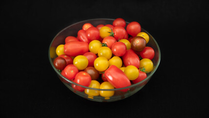 
Small cocktail tomatoes, multicolored, round and oval, in a transparent glass container, on black background, close-up	