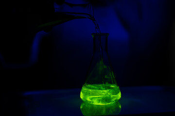 A researcher working with a green fluorescent solution in a glass conical flask in dark biotech laboratory for drug
