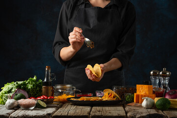 Close-up view of the professional chef in black apron puts the meat filling into the corn tortilla on dark blue background. Backstage of preparing tasty traditional mexican tacos.