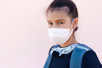 Portrait of teenage girl after school, wearing medical face mask against coronavirus or covid-19. Background of pastel pink color.