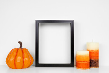 Mock up black frame with fall candles and pumpkin decor on a white shelf. Autumn concept. Portrait frame against a white wall.