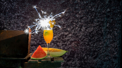 Orange cocktail illuminated by a firework candle and accompanied by pieces of melon and watermelon