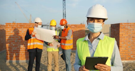 Portrait of Caucasian woman constructor in casque and medical mask standing outdoors at...