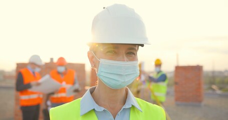Portrait Caucasian young female constructor in casque and medical mask standing outdoor at construction. Close up of female builder at building in helmet. Coronavirus. Builders working on background.