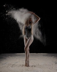 Obraz na płótnie Canvas Beautiful sensual fit slim dancer in black body with long blond hair throwing dust, flour, powder on black background.