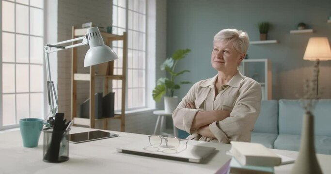 Joyful senior caucasian woman smiling after finishing work at home. Positive tutor leaning back in chair and relaxing while working at home 4k footage