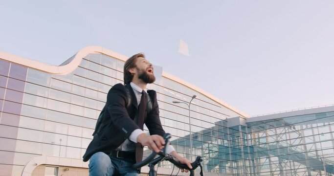 Front view of attractive happy smiling young bearded businessman which riding a bike near modern urban glass construction and throwing business papers over his head