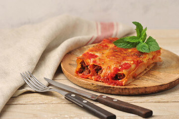 Cannelloni with minced meat, tomato sauce and grated cheese.  Cannelloni on a wooden plate.  Nearby are cutlery and napkin.  Close-up.  Light wooden background.