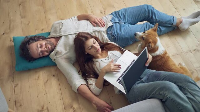 A Lovely Couple Lying on the Floor Using a Laptop, spouses enjoying of Each Other. Near a Couple Lying A Cute Dog. Happy Young Married Couple Talking.