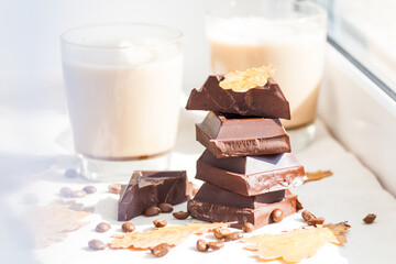 Pieces of dark chocolate, coffee beans and cups of cocoa or coffee with milk on white background. Autumn concept. Sunny day, yellow dry leaves.
