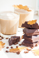 Pieces of dark chocolate, coffee beans and cups of cocoa or coffee with milk on white background. Autumn concept. Sunny day, yellow dry leaves.