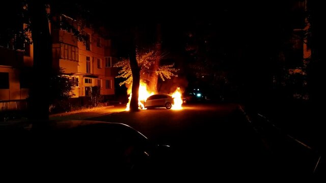 Car burns near a residential building in the light of a special police signal. Theme of vandalism and hooliganism at night. Automobile engulfed in flames of fire. Trees close to burning sedan.