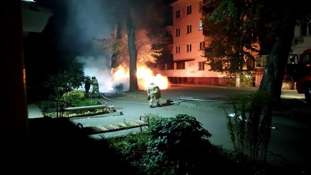 Team of firefighters extinguishes a car burning in a residential area. Theme of vandalism and hooliganism at night. Automobile engulfed in flames of fire. Hand-held shooting from a mobile phone.
