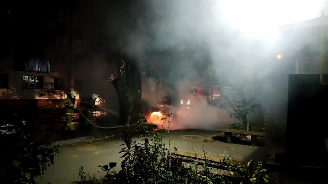 Two firefighters try to put out a burning car at night in a residential area. Theme of vandalism and hooliganism at night. Automobile engulfed in flames of fire. Hand-held shooting from a mobile phone