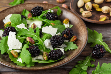 Fresh salad with arugula, feta cheese, blackberries and pistachios in a plate on rustic wooden background